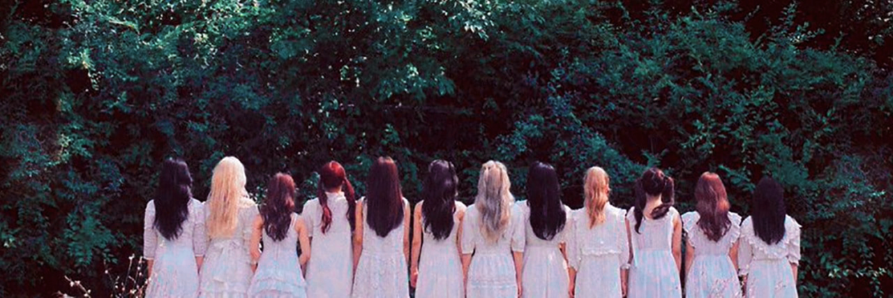 One of the concept photos from ++ era. The members of LOONA are standing with their backs to the viewer in front of a forest. They are wearing white dresses.