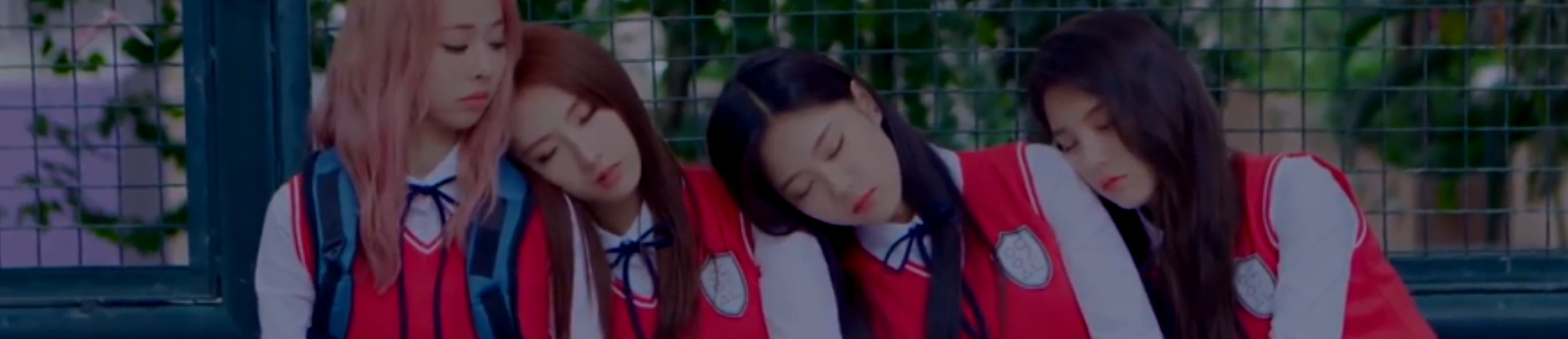 A screenshot from the You and Me Together music video. The members of Loona 1/3 are all sitting on a bench together. From left to right: Vivi, Haseul, Hyunjin, Heejin. Vivi is awake, turned to the right looking at the others. The other three are asleep, with their heads rested on each other's shoulders in a sort of domino effect. They are all wearing their red school uniforms. Vivi is wearing a bulky backpack. Vivi has long light pink hair, Haseul has long brown hair, and Hyunjin and Heejin both have long black hair. They are sat in front of a blue fence, behind which are blurry trees.