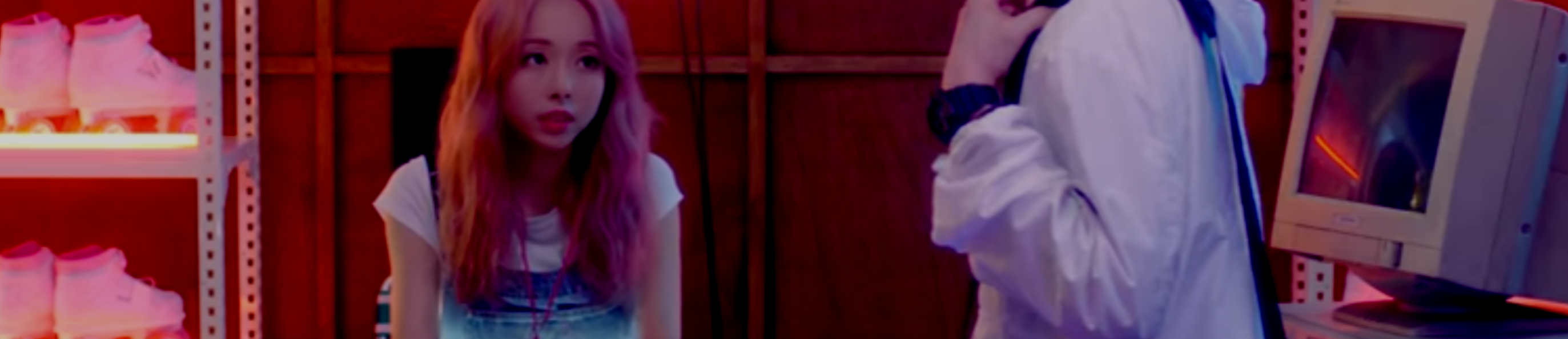 A cropped screenshot from the Everyday I Love You music video. Vivi is sitting behind a desk, wearing a white shirt and blue overalls. She has long light pink hair. She is looking up at a boy who has just entered the room, whose face has been cropped out. Behind Vivi is a rack of roller skates, with neon lights lining the shelves. There is a old looking computer on the right sitting on the desk. The wall behind them is brown and wooden.