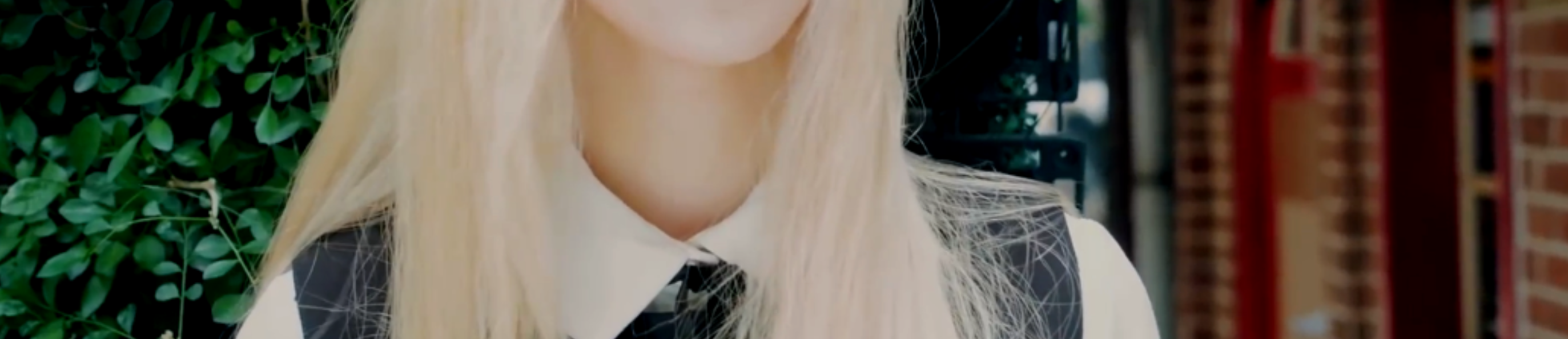 A cropped screenshot of the Everyday I Need You music video. Jinsoul is standing in front of green leaves, with a brick wall in the background. She has long blonde hair and is wearing a black vest over a white button up shirt. However, the image has been cropped so her face isn't visiible.