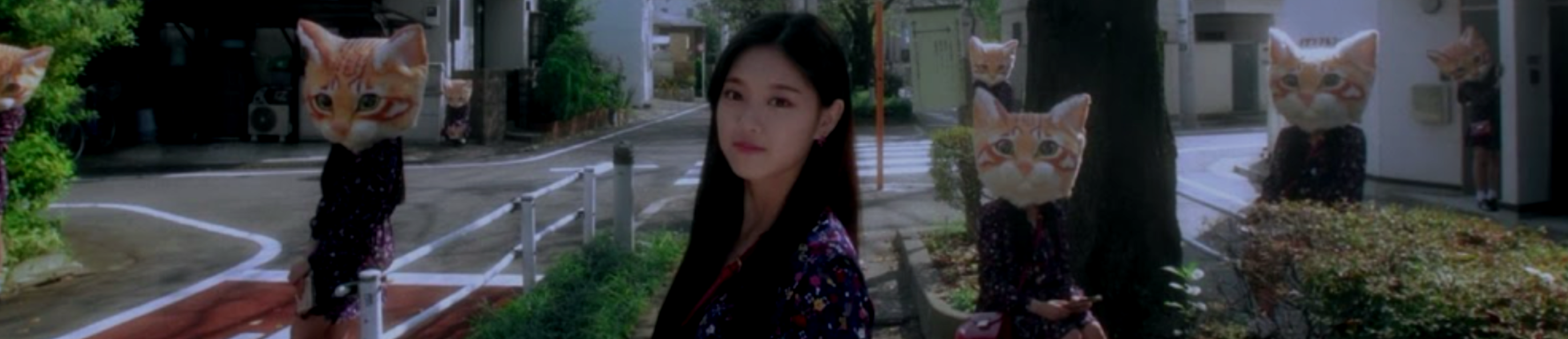 A cropped screenshot from the Around You music video. Hyunjin is standing on a street, wearing a purple dress. She has long black hair. There are four clones of her, wearing the same purple dress and a large orange cat head, spread out across the street behind her.