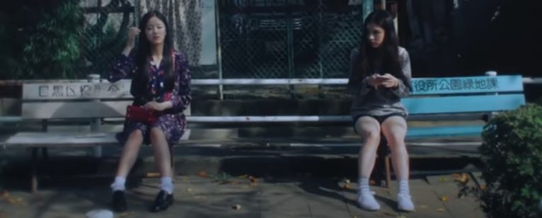 Hyunjin and Heejin sitting on benches. Heejin is looking over at Hyunjin, who is not wearing the cat head.