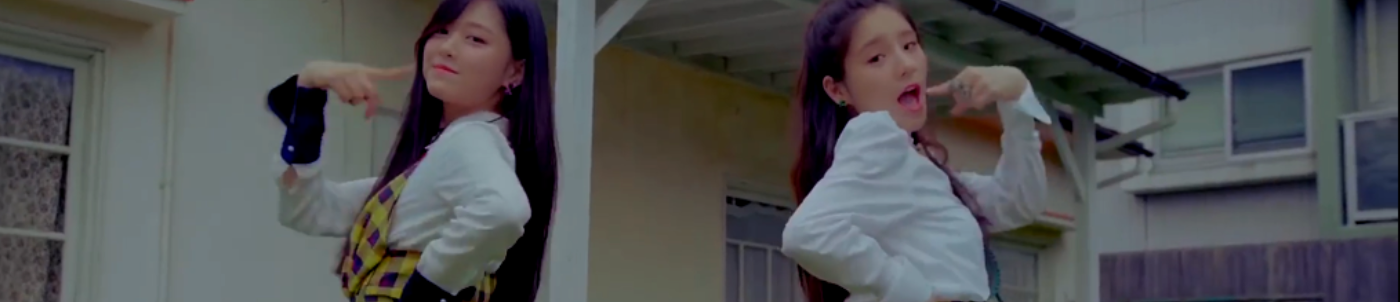 A cropped screenshot from the I'll Be There music video. Hyunjin and Heejin are dancing back to back in front of a house, with their pointer fingers poking their cheeks. Hyunjin is on the left wearing a white long sleeve shirt with a yellow plaid sleeveless dress over it. She has long black hair. Heejin is on the right wearing a white longe sleeve shirt. She has long dark brown hair.