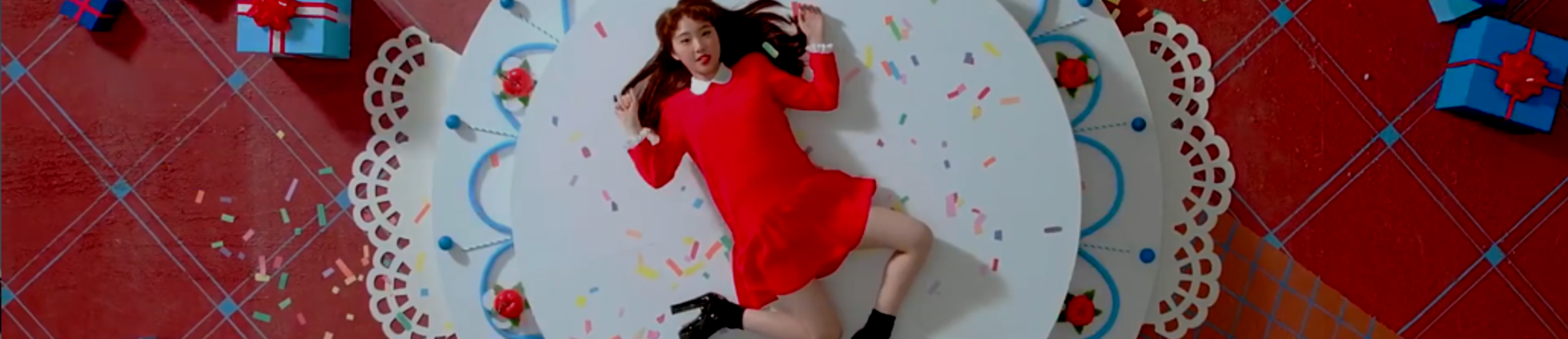 A cropped screenshot from the Kiss Later music video. Yeojin is laying on a large white podium, looking up at the viewer. Her arms and legs are awkwardly splayed out around her. She is wearing a short red dress with a white collar, and heeled black boots. She has long reddish brown hair. The podium has multicolored sprinkles on top of it, with blue stripes and red stairs on the edges. White lace surrounds the podium, and there are blue and red gift boxes on the ground.