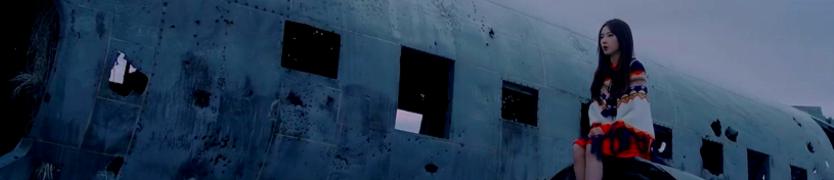 A cropped screeshot from the Let Me In music video. Haseul is sitting on the wing of the crashed plane. She is wearing a intricately patterned shawl. She has long brown hair. The sky behind the plane is gray.