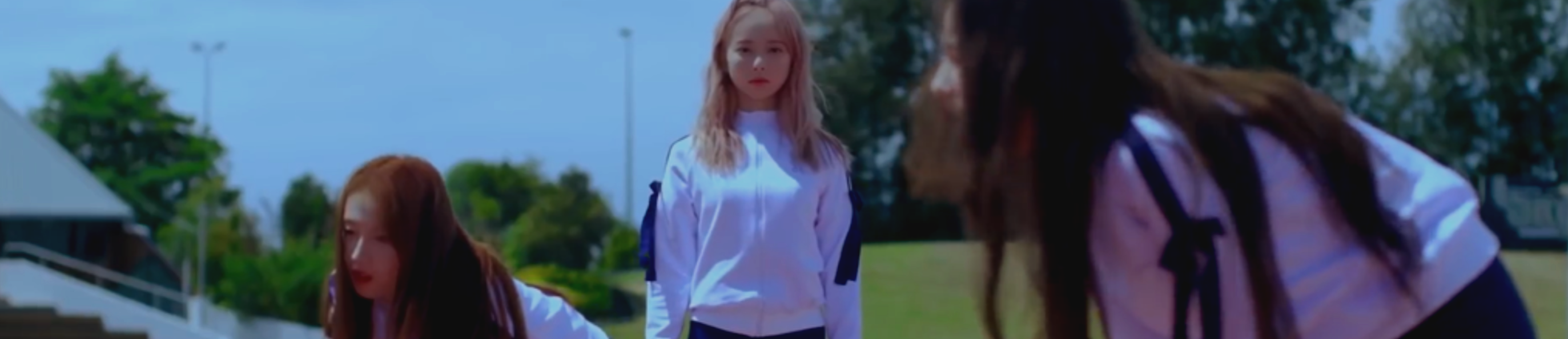 A cropped screenshot from the Love and Live music video. Vivi is standing in the middle in her white and navy track uniform, perfectly stoic. She has long light pink hair. In front of her is Haseul, also in her track uniform. In contrast, she is bent over catching her breath, clearly exhausted. She has long brown hair. In the foreground is Heejin, also in her track uniform and bent over catching her breath. She has long dark brown hair. Behind them are bleachers and bright green grass, with trees in the background. The sky is a bright blue.