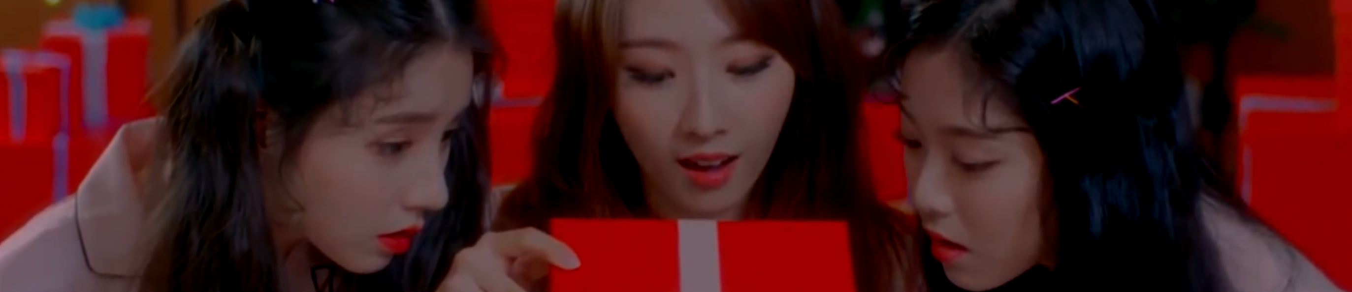 A cropped screenshot from The Carol music video. From left to right, Heejin, Haseul, and Hyunjin are sitting wearing pajamas. Heejin has long dark brown hair styled into two large pigtails. Haseul has long brown hair. Hyunjin has long black hair. They are all looking at a bright red gift box as Haseul begins to open it. The background is full of stacks of the same bright red presents.