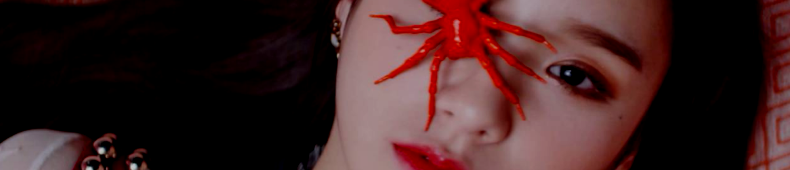 A cropped screenshot from the Vivid music video. Heejin is laying on a red patterned floor, staring up at the viewer. There is a red spider prop covering her right eye. She has long dark brown hair.