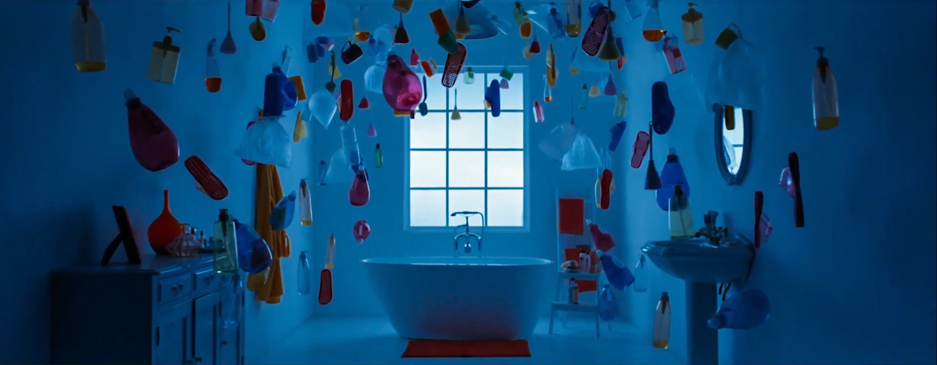 A screenshot of a bathroom. There is a bathtub in the middle, with a square window above it. On the left is a drawer with various items on it, and on the right is a sink with an oval-shaped mirror above it. Floating all around the room are various multi-colored objects. The entire scene is lit by dim blue lighting.