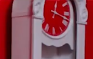 A cropped screenshot of a white clock with a red face, against a red wall. It appears to read around 12:18.