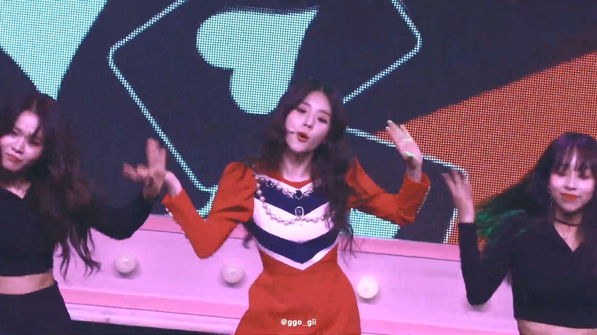 An image of Heejin performing Vivid live. She is wearing a red dress and dancing, with backup dancers wearing black surrounding her. Behind her is an LED screen, which is displaying a video of playing cards falling.