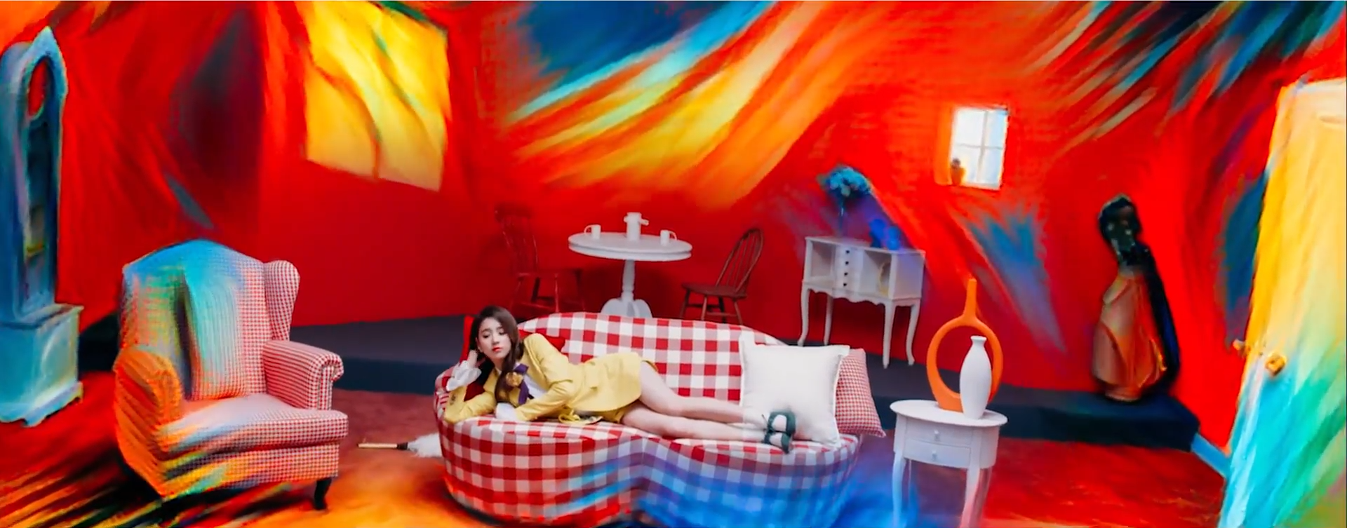 A screenshot of Heejin. She is in her yellow outfit, laying on a red and white plaid couch in the middle of a red room. A visual effect is creeping in from the edges of the screen, with transforms the image into a colorful painting.