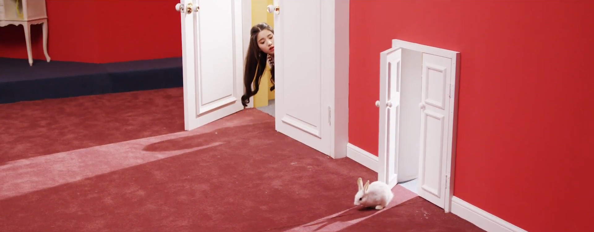 A screenshot of Heejin. She is peaking out from behind a white door, with only her head being visible. She is looking down at a white rabbit, which is emerging from a smaller white door.