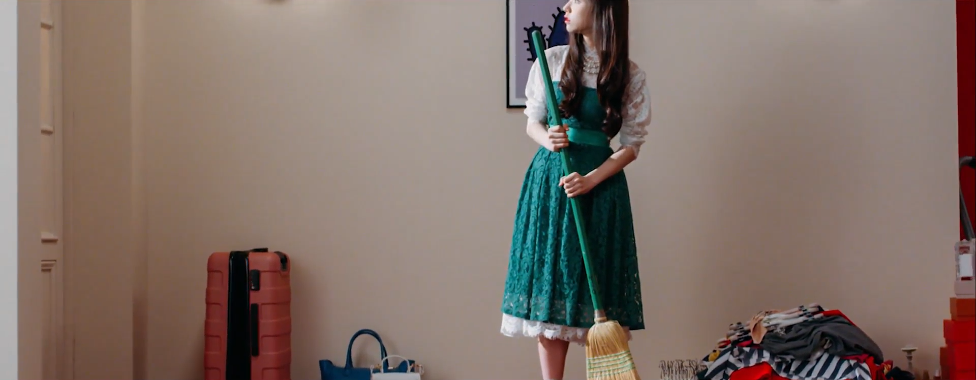 A screenshot of Heejin wearing the green dress. She is holding a broom, as if in the process of sweeping. She is looking towards the left, where a light is shining on her. There is a suitcase and piles of clothes on the floor around her.