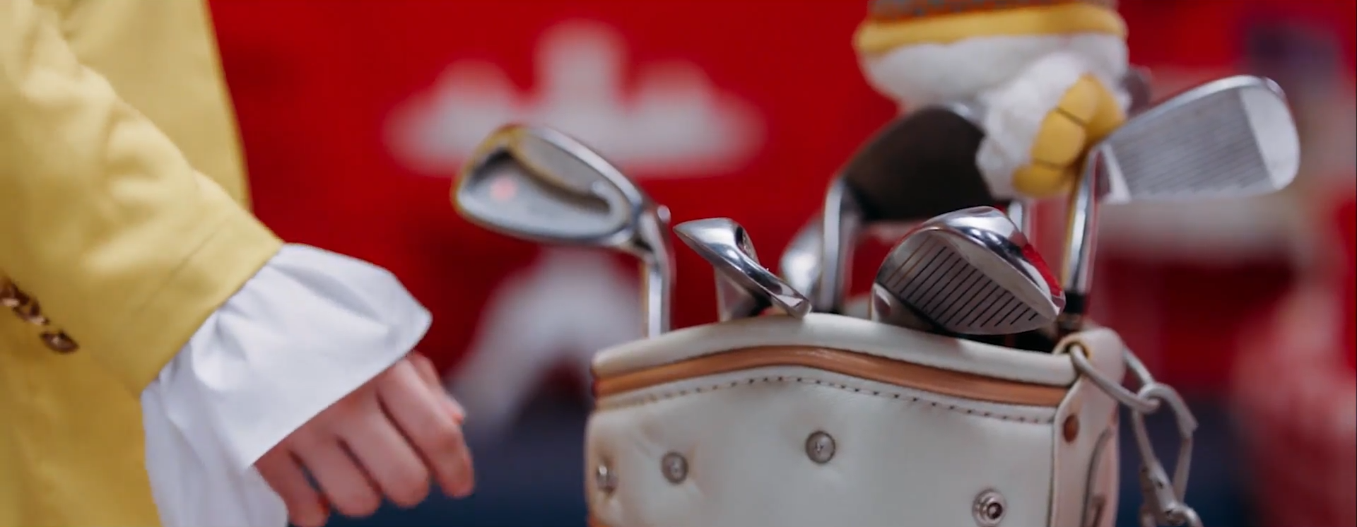 A screenshot of Heejin reaching her arm out to a bag full of multiple golf clubs. She is wearing her yellow outfit. The image is cropped so only her arm and a bit of her torso can be seen.