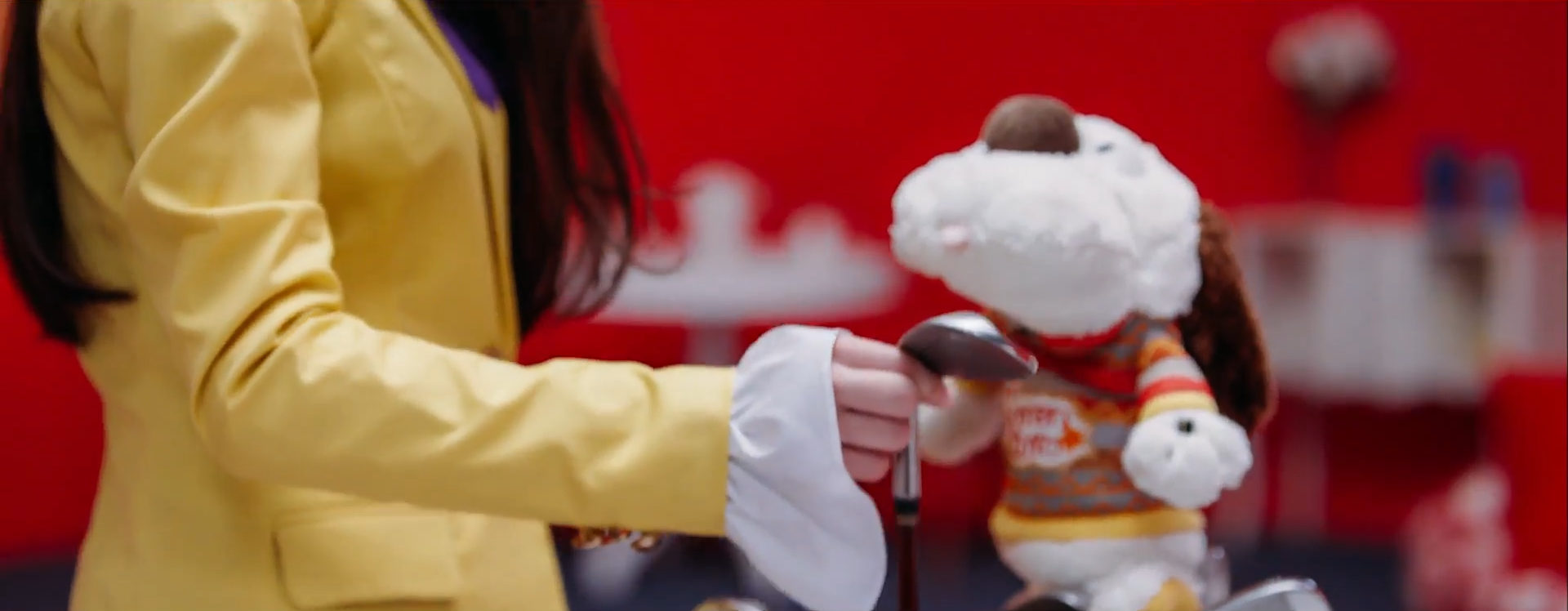 A screenshot of Heejin, immediately following the preivous one. She is grabbing one of the golf clubs, pulling it up out of the bag. There is a dog stuffed animal on top of the bag, wearing a shirt. The shot has panned upward a bit since the previous screenshot, so now more of Heejin's torso and some of her hair can be seen.