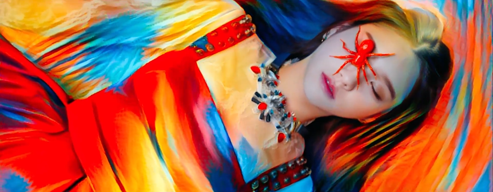 A screenshot from the Vivid music video. Heejin is laying on the floor with her eyes closed. She is wearing a red dress, and there is a red fake spider over her right eye. The entire image has an effect over it that makes it appear like a colorful painting.