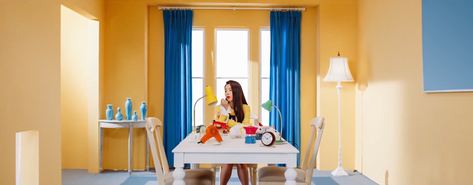 A screenshot of Heejin. She is sitting at the end of a table with various items on it. The walls are yellow, and she is wearing her yellow outfit. She has one elbow resting on the table, with her head tilted to the left as she sings. There is a large window behind her, with blue curtains.