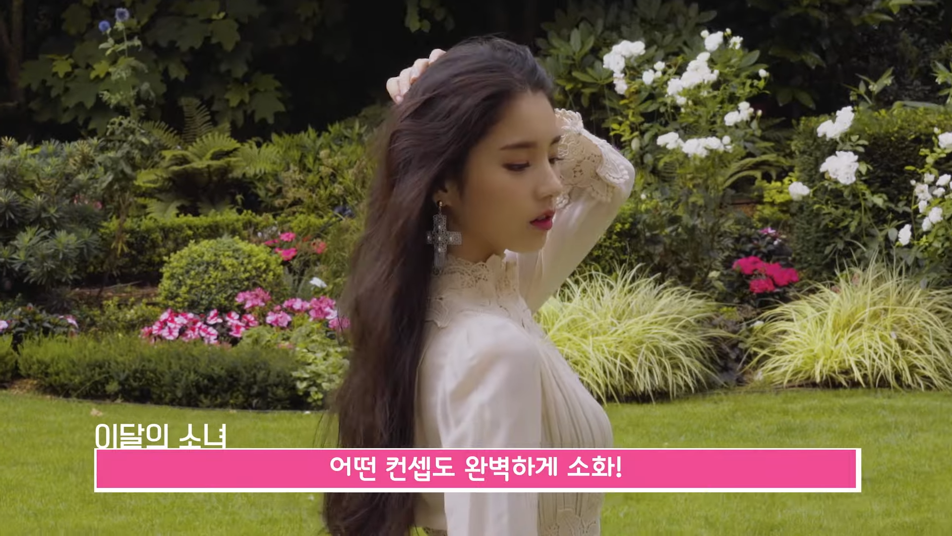 A screenshot from an episode of LOONA TV. Heejin is standing in a lush garden, wearing a lacy peach dress. She is wearing a large ornate cross earring. She is posing, with one arm behind her head.