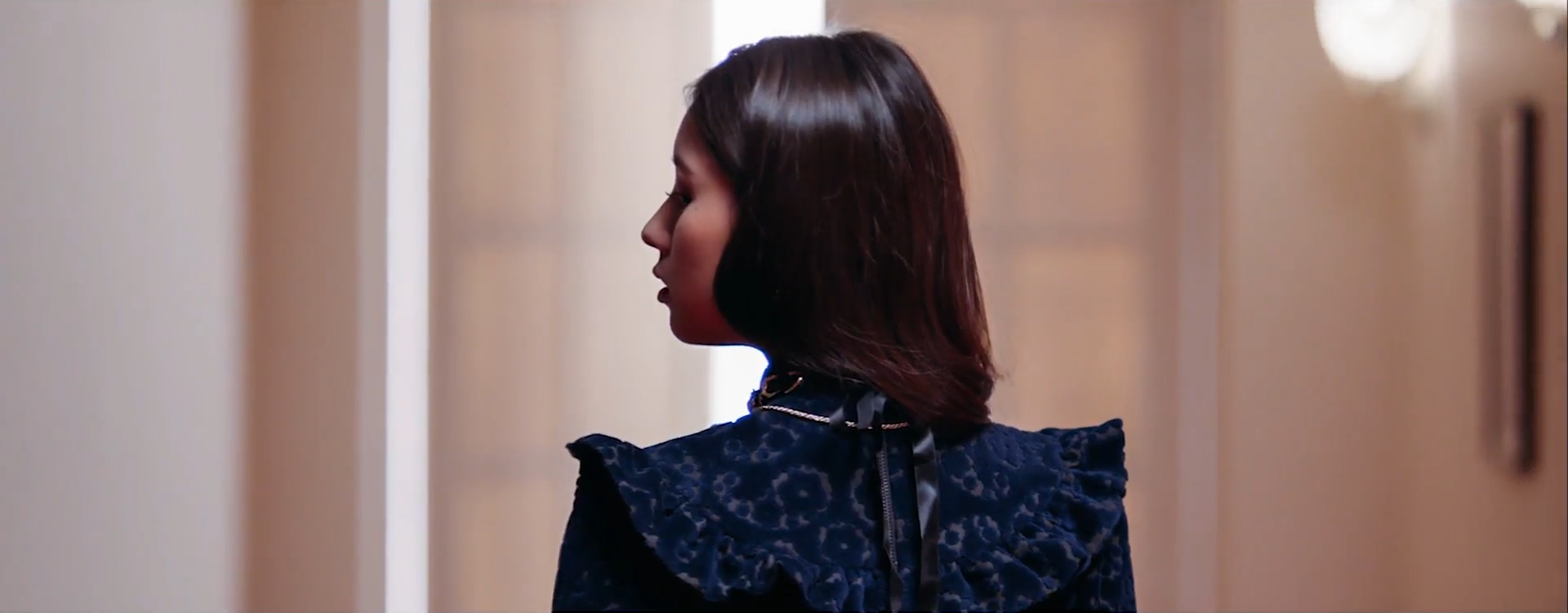 A screenshot of Homeowner Heejin in the black dress. Her back is to the viewer, with her head tilted to left so that you can see her face. The background is the door she was shown in front of before.