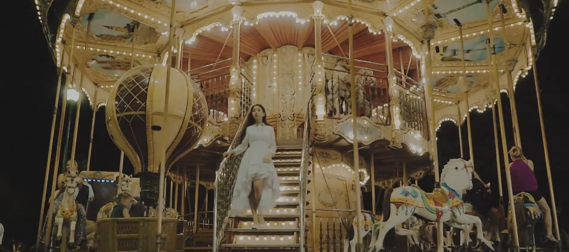 A screenshot of Heejin wearing a dress, standing on the staircase leading up to a golden carousel.