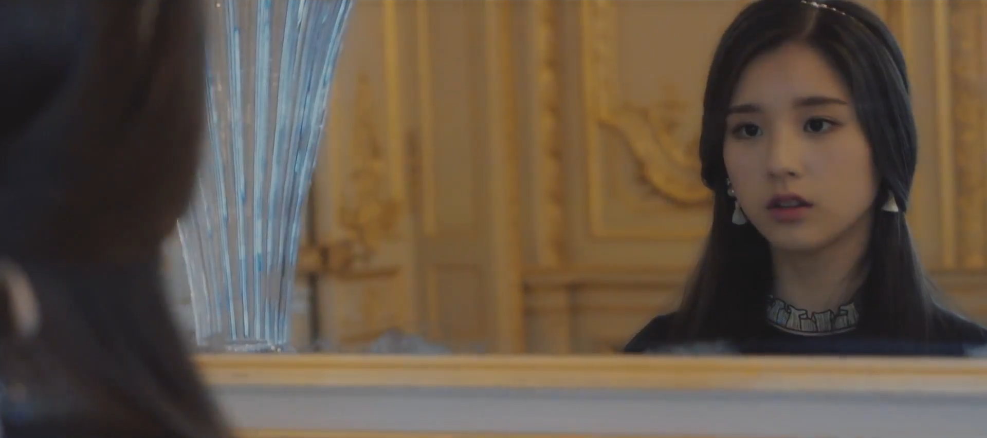 A screenshot of Heejin standing in a golden room. She is staring at her reflection in a mirror, with a blank, somewhat surprised expression.