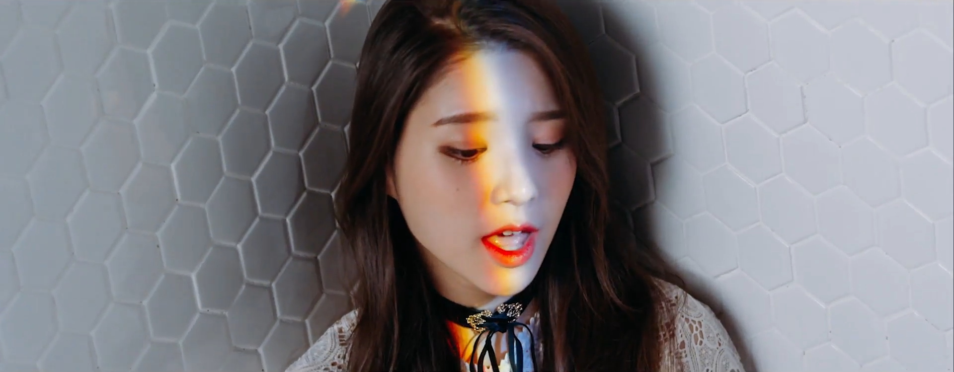 A screenshot of Heejin. She is standing against a white wall with a hexagonal pattern. She is looking downwards, in the middle of singing with her mouth open. There is a rainbow shining across her face.