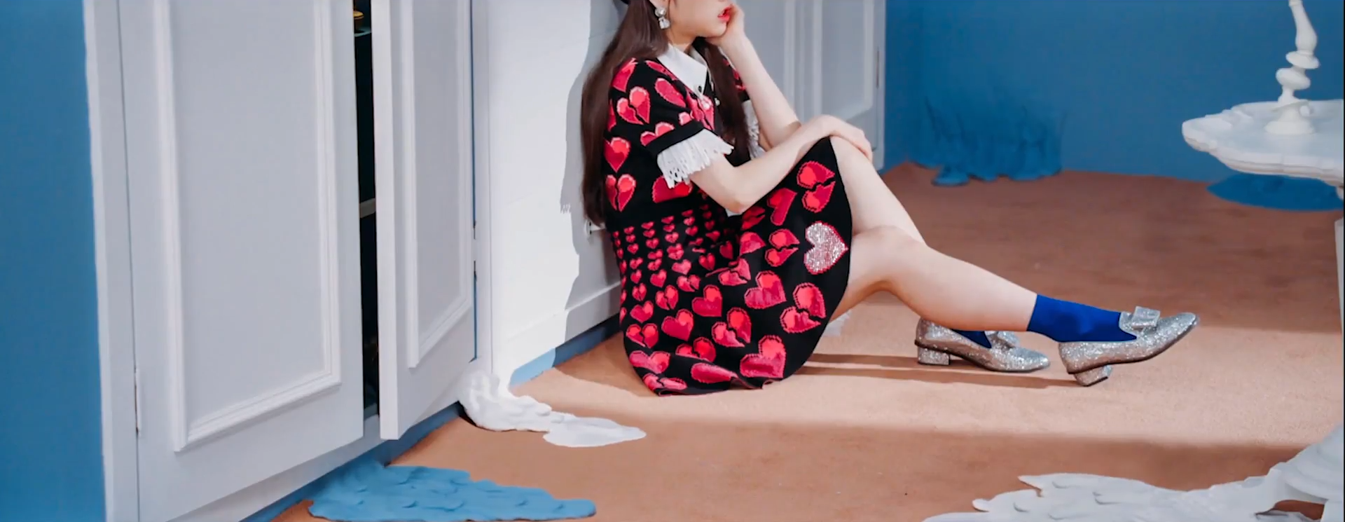 A screenshot of Heejin. She is wearing a black dress with a pattern of pink whole and broken hearts covering it. She is wearing blue socks with glittery silver shoes. She is sitting against a white door, with her head resting in her palm. There is a straight line of light illuminating her, seemingly from an open door. Around her, the walls and furniture are melting into a kind of goo.