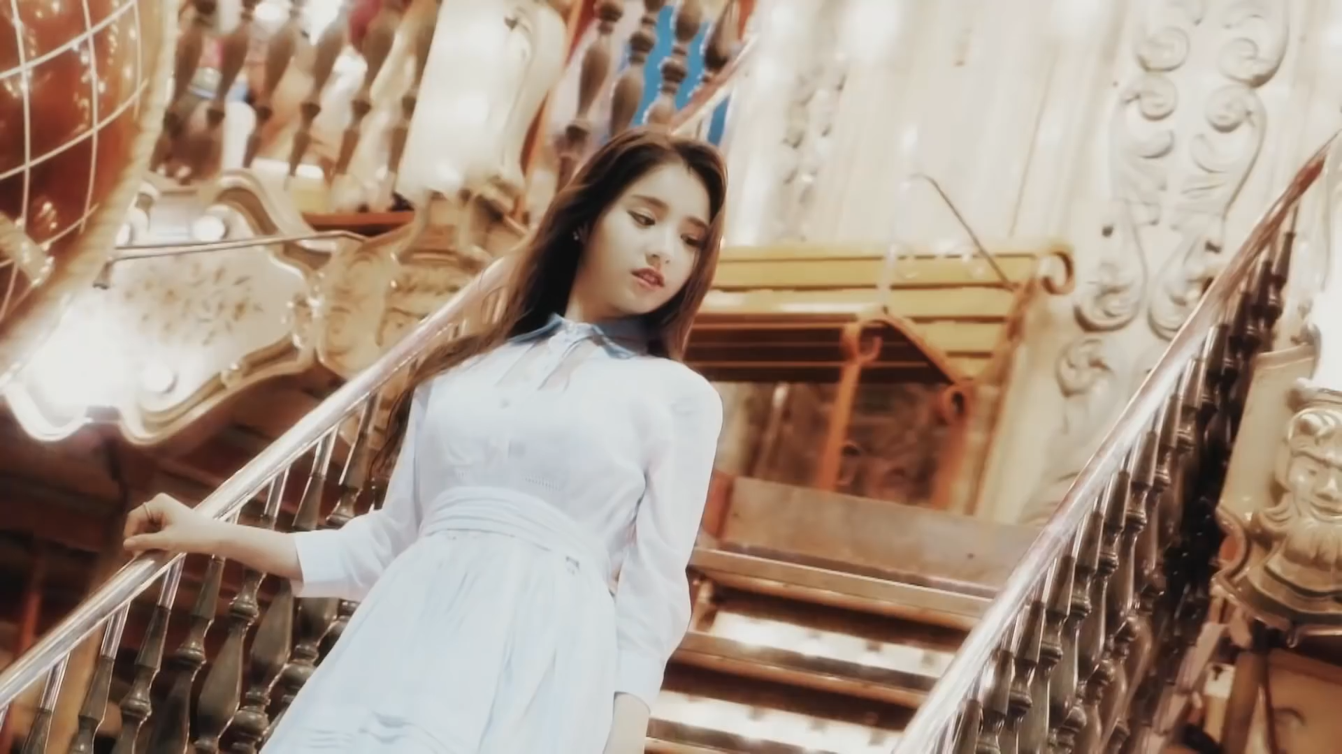 A screenshot from '[Teaser] LOONA's 1st Member HeeJin Seoul Teaser.' She is wearing a dress and standing on a staircase. She is facing the left, with her head tilted to look at the right. She's got a sort of half smile.