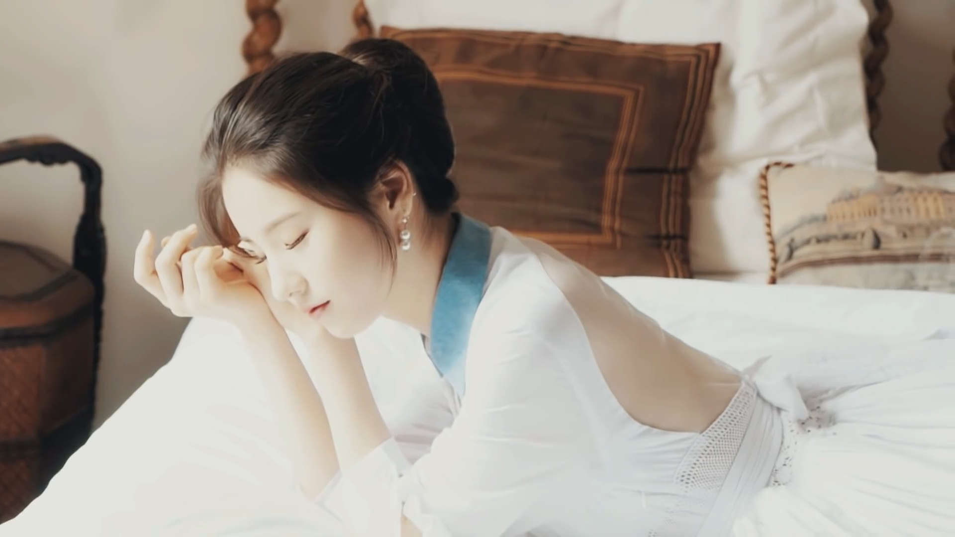 A screenshot from '[Teaser] LOONA's 1st Member HeeJin Seoul Teaser.' She is wearing a dress and laying down on her stomach on a bed. Her head is resting on her hands, and her eyes are closed as if she's asleep.