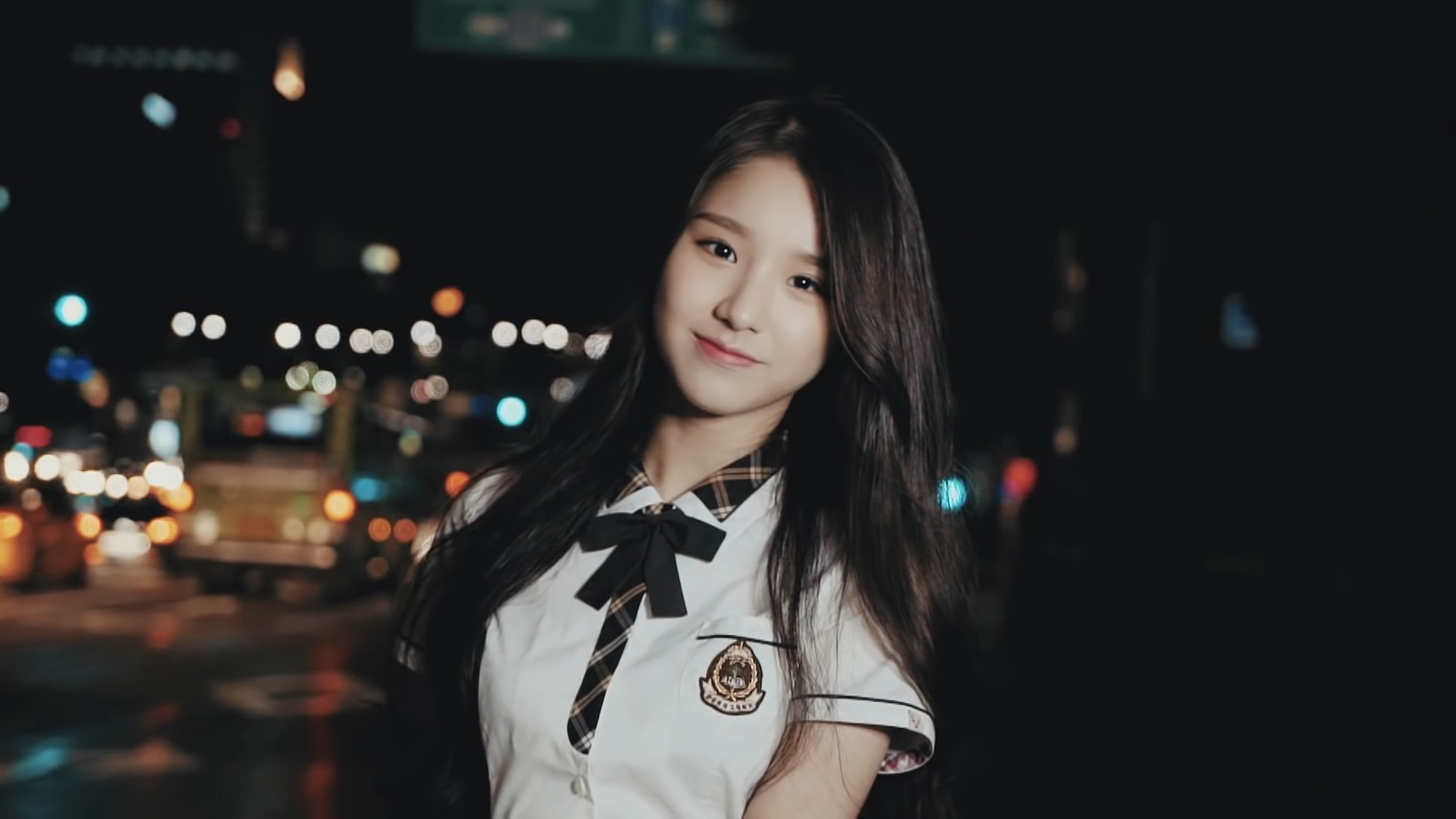 A screenshot from '[Teaser] LOONA's 1st Member HeeJin Seoul Teaser.' Heejin is looking at the viewer and smiling in front of a blurry cityscape. She is wearing a schoolgirl uniform.