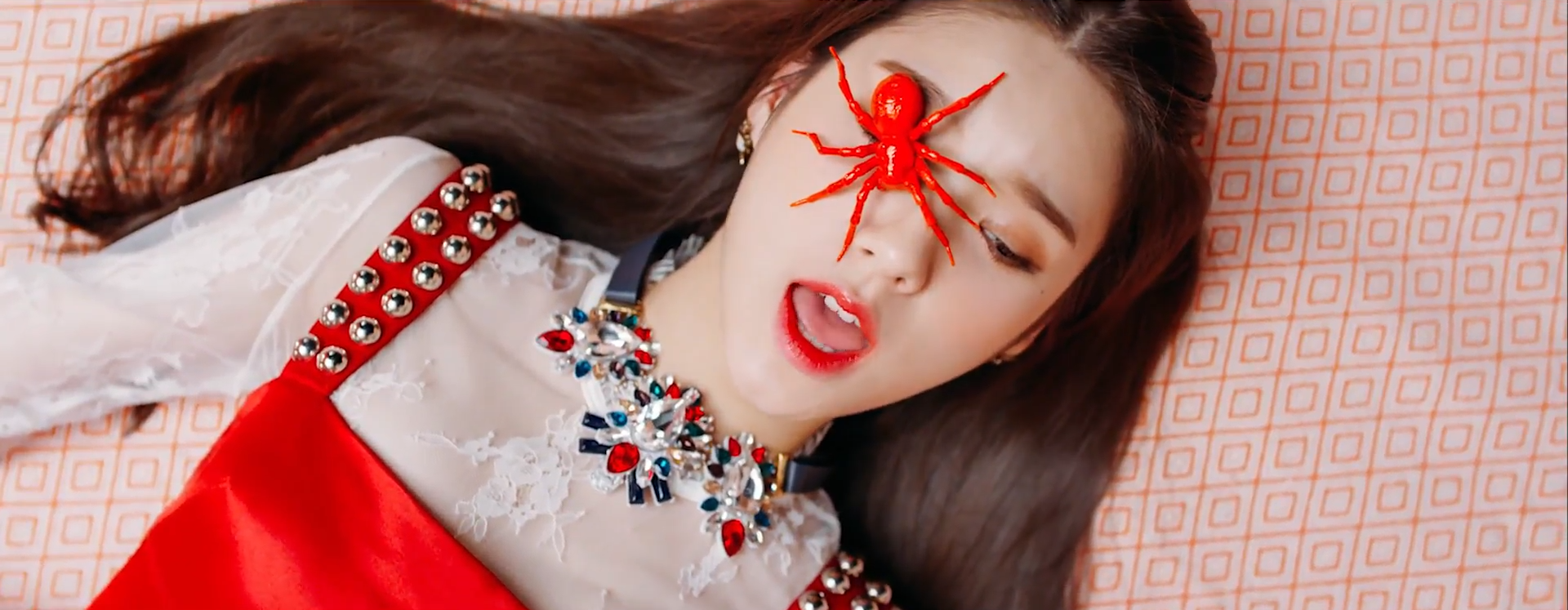 A screenshot of Heejin. She is wearing a red dress and laying on the floor. There is a fake red spider on her right eye. She is looking downwards, with her mouth open as she's in the middle of singing.