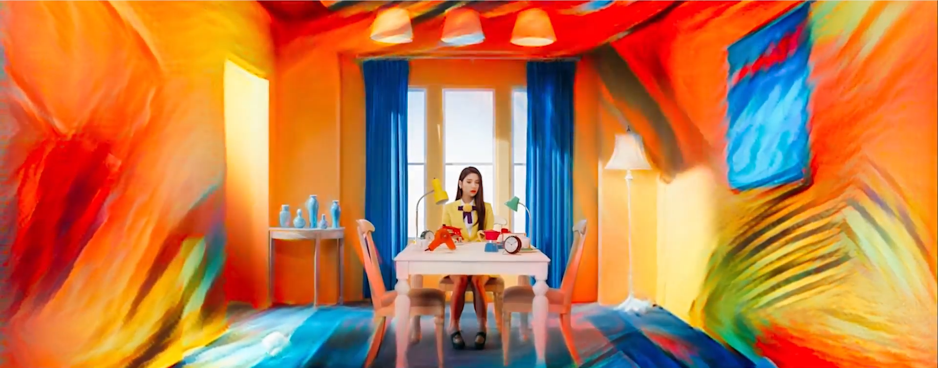 A screenshot of the same room with the table. Heejin is now back in her chair, wearing her yellow outfit. The painting effect is coming in from the edge of the screen.