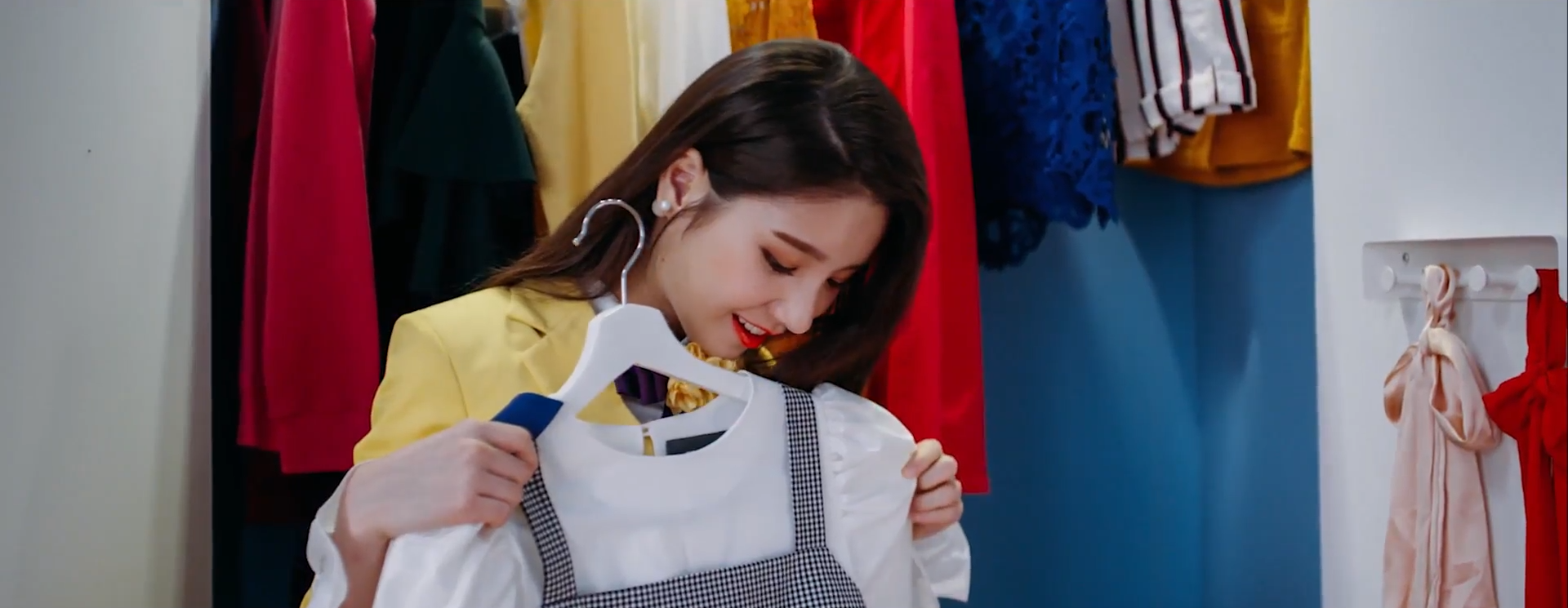 A screenshot of Heejin. She is in the same spot as the previous image, only now she's holding a dress on a hanger out in front of her. She is looking down at it with a smile.