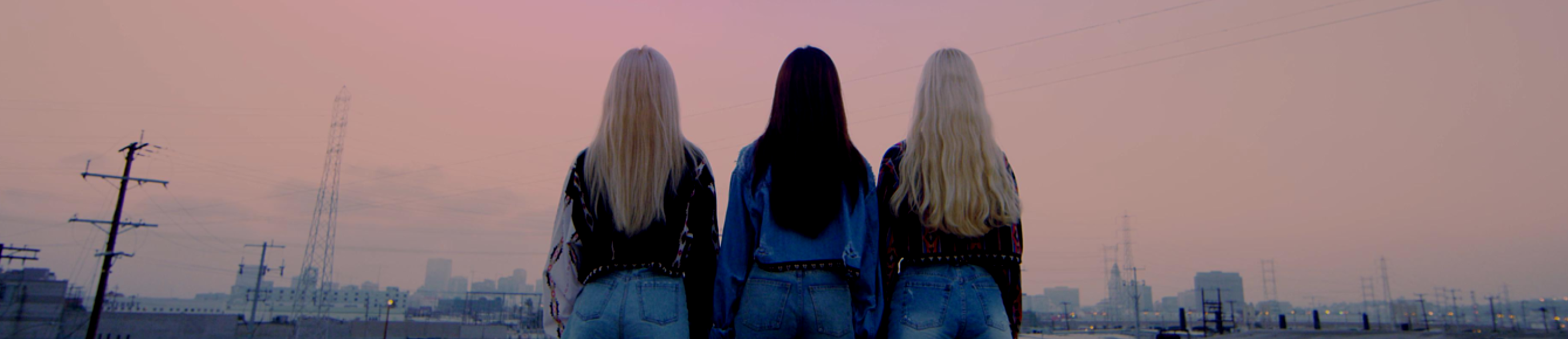 A cropped screenshot from the Girl Front music video. The members of Odd Eye Circle are standing with their backs to the viewer. From left to right: Kim Lip, Choerry, Jinsoul. They are wearing patterned denim jackets and blue denim shorts. Kim Lip and Jinsoul both have long blonde hair, while Choerry has long dark brown hair. The sky in front of them is a hazzy pinkish color. The silhouette of a cityscape is visible in front of them.