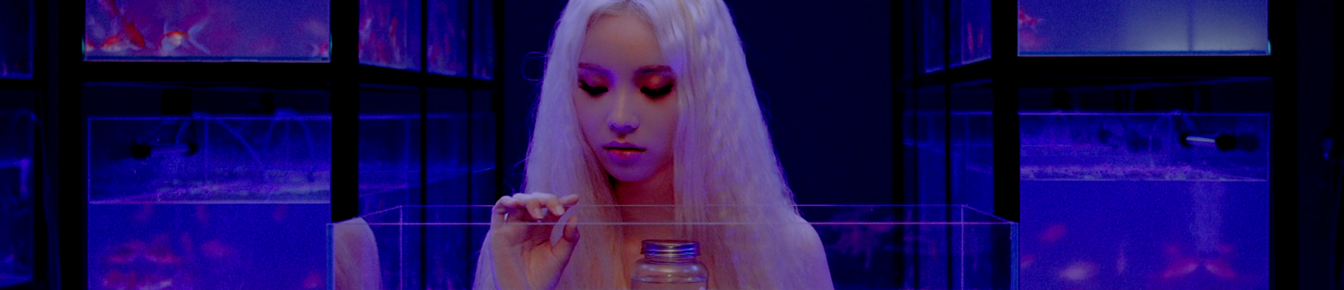 A cropped screenshot from the Singing in the Rain music video. Jinsoul is standing in front of a fish tank, holding a glass jar full of water. She has long blonde hair and is wearing a blue dress. There are rows of other blue fish tanks behind her.