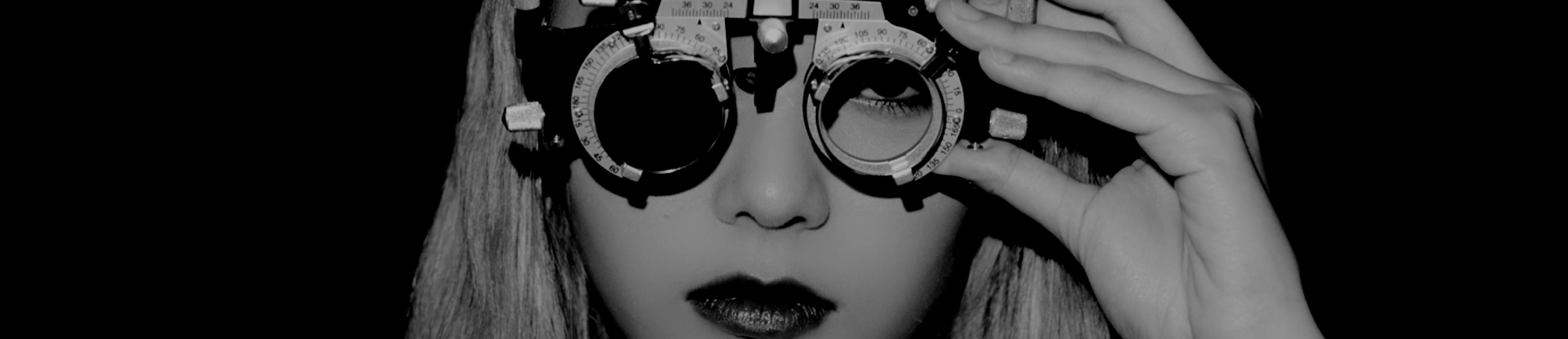 A cropped screenshot from the Sweet Crazy Love music video. Jinsoul is staring at the viewer through the lenses of a phoropter, the machine using to assess one's eyesight. Her right eye is blocked, and she has a flat expression. She has long blonde hair. The background is entirely black, and the whole image is in black and white.