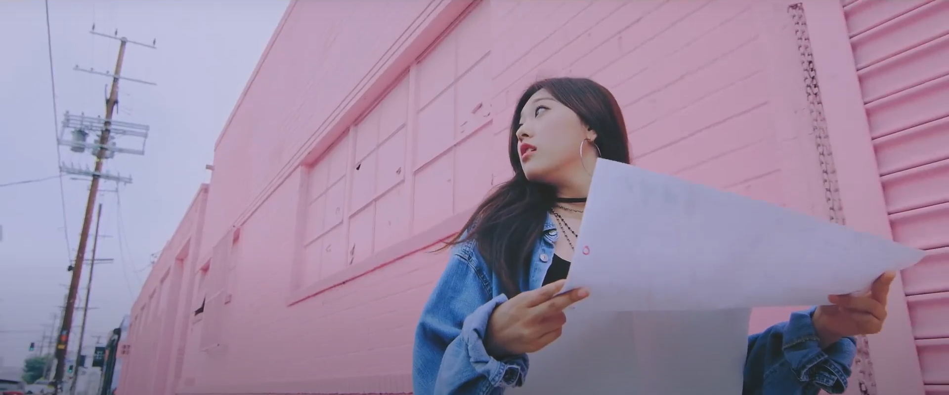 A cropped screenshot from the Girl Front music video. Choerry is holding a white paper map in front of her and looking around to the left. She has long brown hair and is wearing a blue denim jacket, a black shirt, and large hoop earrings. She is walking in front of a light pink wall, and there is a powerline behind her. The sky is a clear gray.