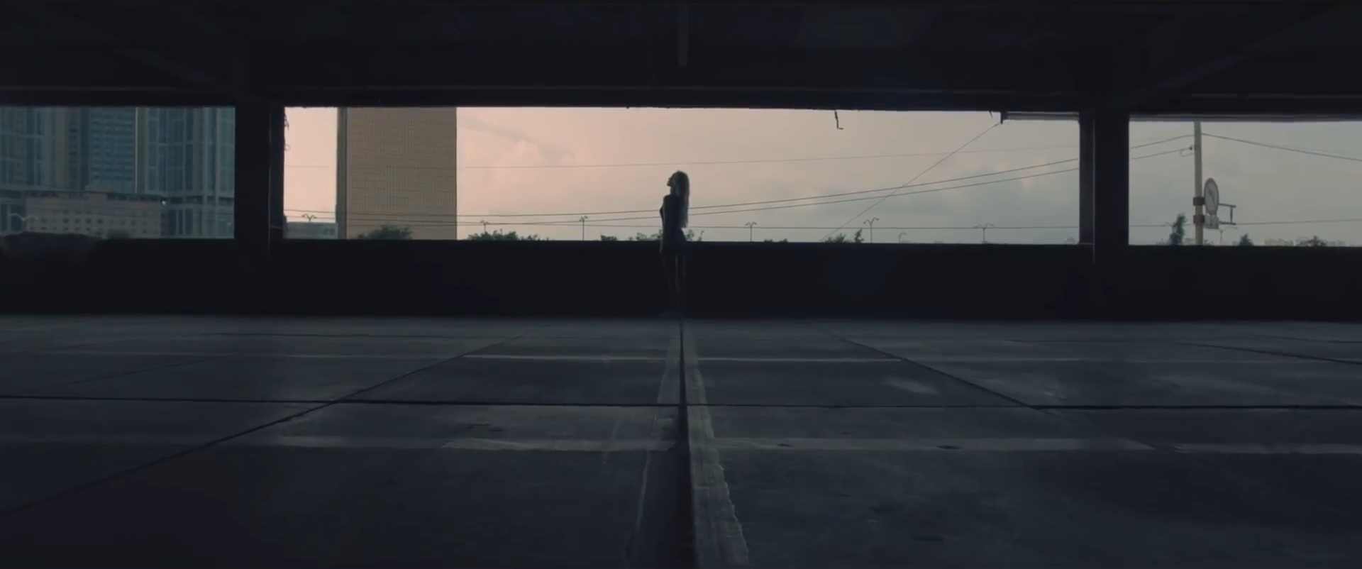 A screenshot from the Hi High music video. The silhouette of Jinsoul is standing looking up at the sky. The background is silhouettes of buildings around her in front of a gray cloudy sky.