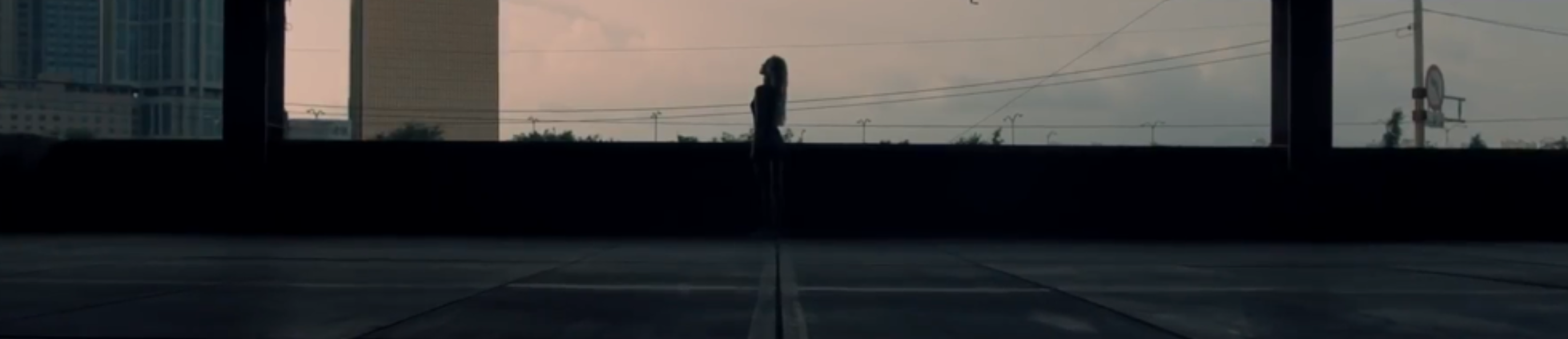 A screenshot from the Hi High music video. The silhouette of Jinsoul is standing looking up at the sky. The background is silhouettes of buildings around her in front of a gray cloudy sky.