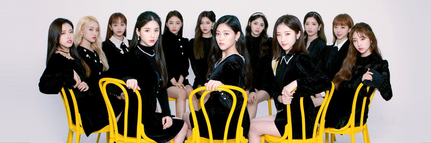 A cropped image of the members of Loona, from the official Japanese website. The members are all wearing black and white dresses and looking out at the viewer. They are sitting in bright yellow chairs in a circle. The background is a white void.