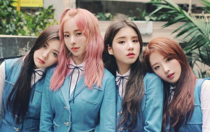 A picture of LOONA 1/3. From left to right: Hyunjin, ViVi, Heejin, Haseul. They are wearing their blue school uniforms. Hyunjin is leaning on ViVi's shoulder, while Haseul is leaning on Heejin's shoulder. Hyunjin and Heejin have long black hair, Vivi has long light pink hair, and Haseul has long brown hair. There is blurry architecture and plants behind them.