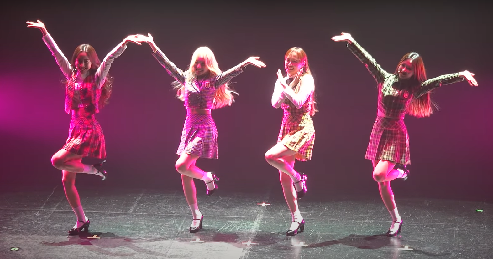 A photo of yyxy performing love4eva at Premier Greeting Meet & Up. From left to right: Yves, Go Won, Chuu, Olivia Hye. They are wearing plaid tops and skirts, with white socks and black shoes. They are on a simple black stage, standing in the middle of a spotlight. Yves, Go Won, and Olivia have their arms in the air with one leg up, making a Y shape. Chuu has one leg up and is crossing her arms in front of her, making a X shape. Altogether, their poses spell out YYXY.