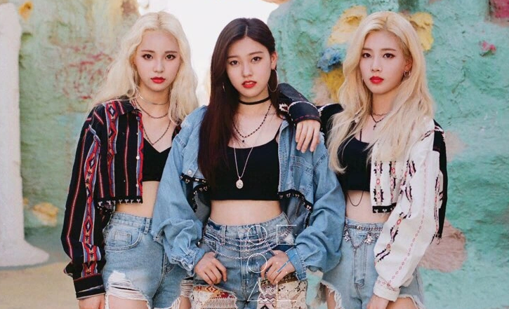 A picture of Odd Eye Circle. From left to right: Jinsoul, Choerry, Kim Lip. They are wearing denim jackets and shorts, with necklaces and hoop earrings. Choerry has her hands on her hips, while Kim Lip rests her right arm on Choerry's shoulder. Kim Lip and Jinsoul both have long blonde hair, while Choerry has long dark brown hair. They are standing in front of oddly colored rock walls with spots.