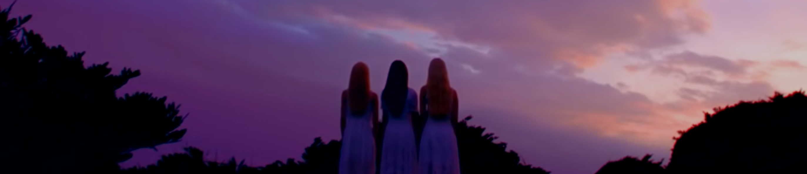 A screenshot from the Reveal teaser. The members of Odd Eye Circle are standing with their backs to the viewer. From left to right: Kim Lip, Choerry, Jinsoul. They are wearing long white dresses. Kim Lip and Jinsoul both have long blonde hair, while Choerry has long dark brown hair. They are surrounded by the silhouettes of bushes. They are looking up at the sky, which is various shades of blue and pink with large fluffy pink clouds. Within the clouds are three shining moons. On the left is a small red moon, in the middle is a large purple moon, and on the right is a medium sized blue moon.