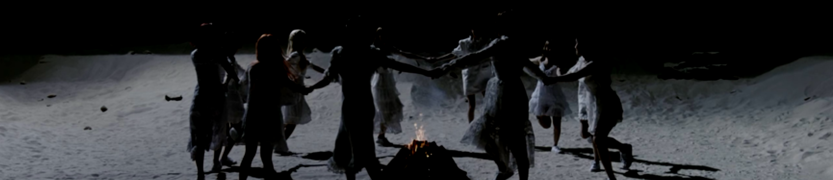 The members of LOONA dancing on the moon around a campfire, from the Why Not music video. They are wearing white dresses and are skipping in a circle, holding hands with each other. They are standing on the sandy gray surface of the moon. Black space is behind them, with the Earth floating nearby.