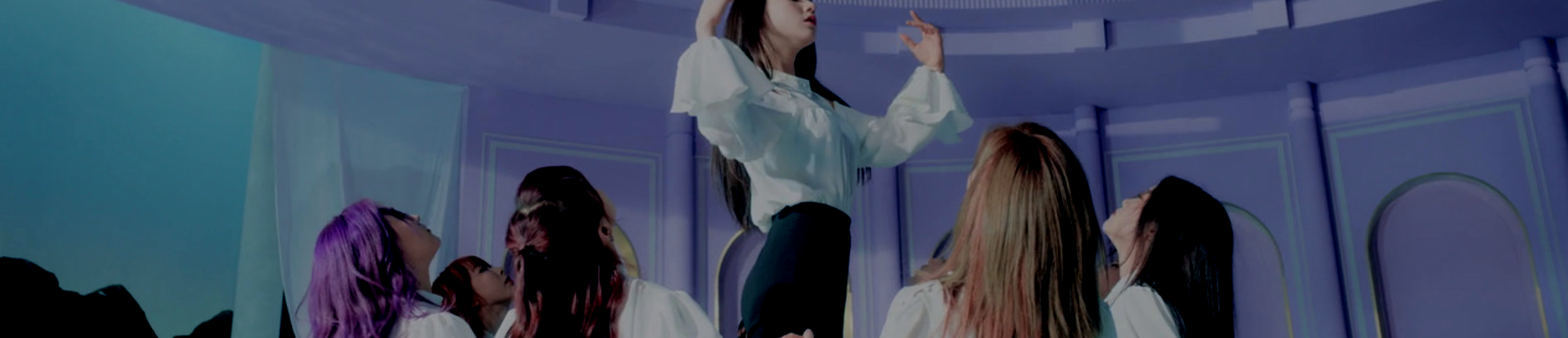 A cropped screenshot from the Butterfly music video. The members of Loona are in the opening formation of the Butterfly choreography, sitting around in a circle with their hands linked as Heejin stands in the middle with her hands raised. They are wearing frilly white shirts and black pants. They are within a large dome, with a white curtain and blue sky behind them.