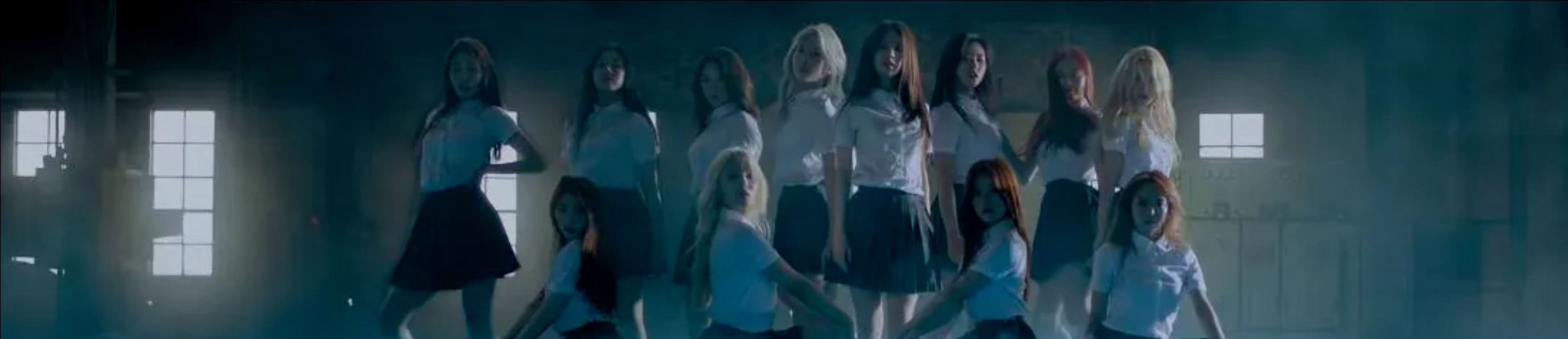 A cropped screenshot from the favorite music video. The members of Loona are posing in an abandoned warehouse. They are all wearing white short-sleeved button-up shirts and navy pleated skirts. They are all looking at the viewer.