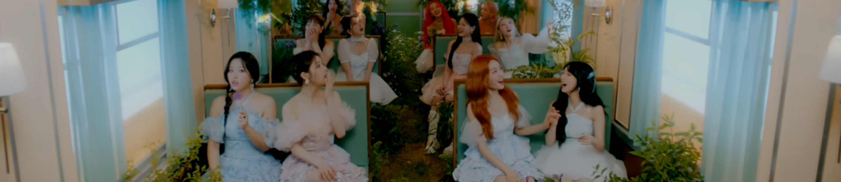 A cropped screenshot from the Flip That music video. The members of Loona are sitting inside a train, wearing lightly colored frilly dresses. They are smiling and looking around, surprised as butterflies fly in the air. The train has teal seats, with white walls and blue curtains on the windows. There are white lamps in the foreground. Green foliage is growing all around the seats.