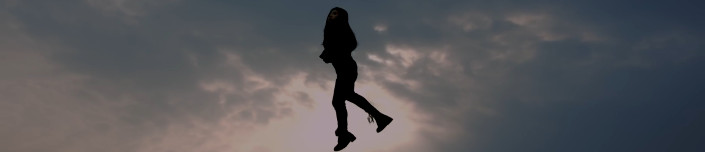 A cropped screenshot from the #1 teaser. Heejin is floating in the sky, wearing her black outfit from Butterfly. It is a frilly black top with black pants and boots. She has long dark brown hair. The sky behind her is gray and cloudy, with the sun shining through the clouds.